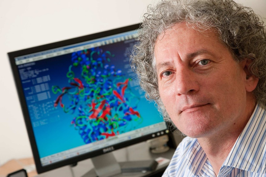 A man looks at a camera with a colourful, complicated graph on a computer behind him.