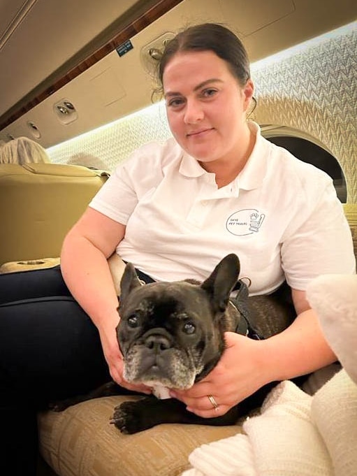 Woman with brown hair and white t-shirt on a small plane holding a small black dog.