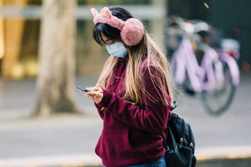 A woman wearing a mask and pink ear muffs looks at her phone.