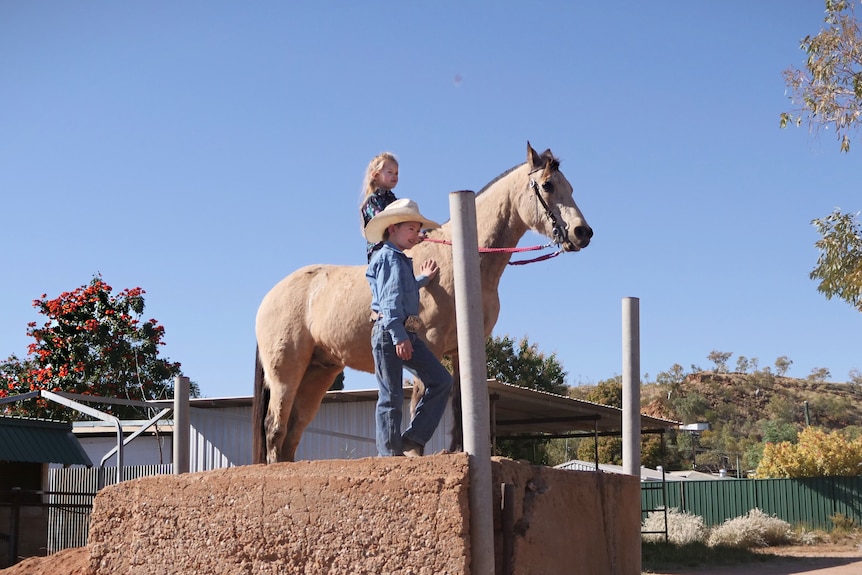 A young girl sits bareback atop her buckskin horse while her brother stands beside them. Both wear full rodeo outfit.