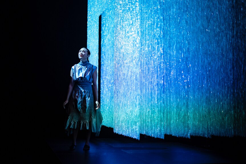 A Chinese Australian in her 30s with long dark hair wears a silver dress and sings onstage in front of blue-lit beaded curtain.