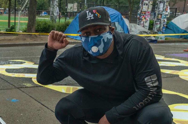 Man in mask, black sweatshirt and pants crouches next to art on the ground, raising right fist