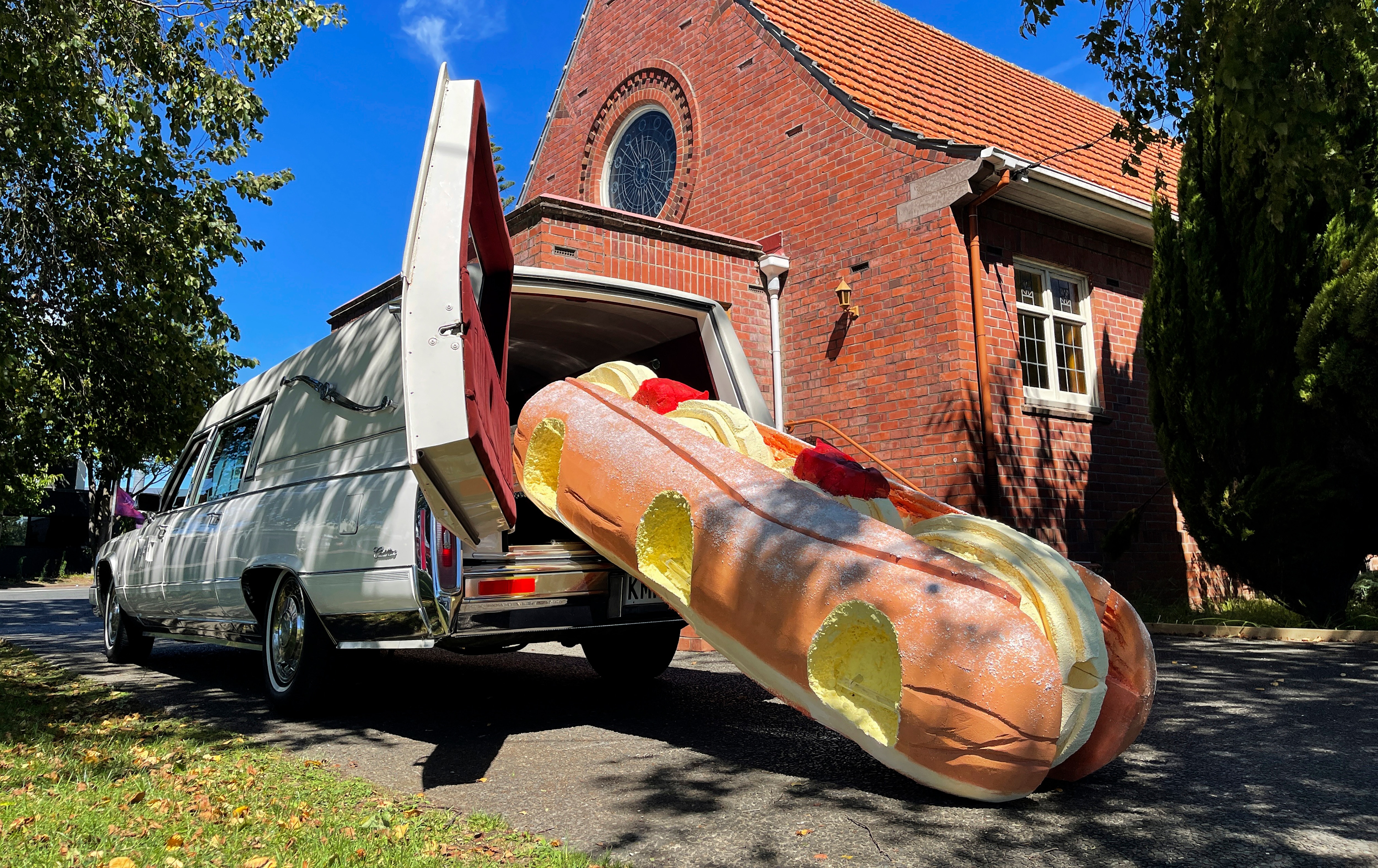 A doughnut-shaped coffin rests halfway out of the boot of a white hearse in front of a church