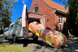 A doughnut-shaped coffin rests halfway out of the boot of a white hearse in front of a church