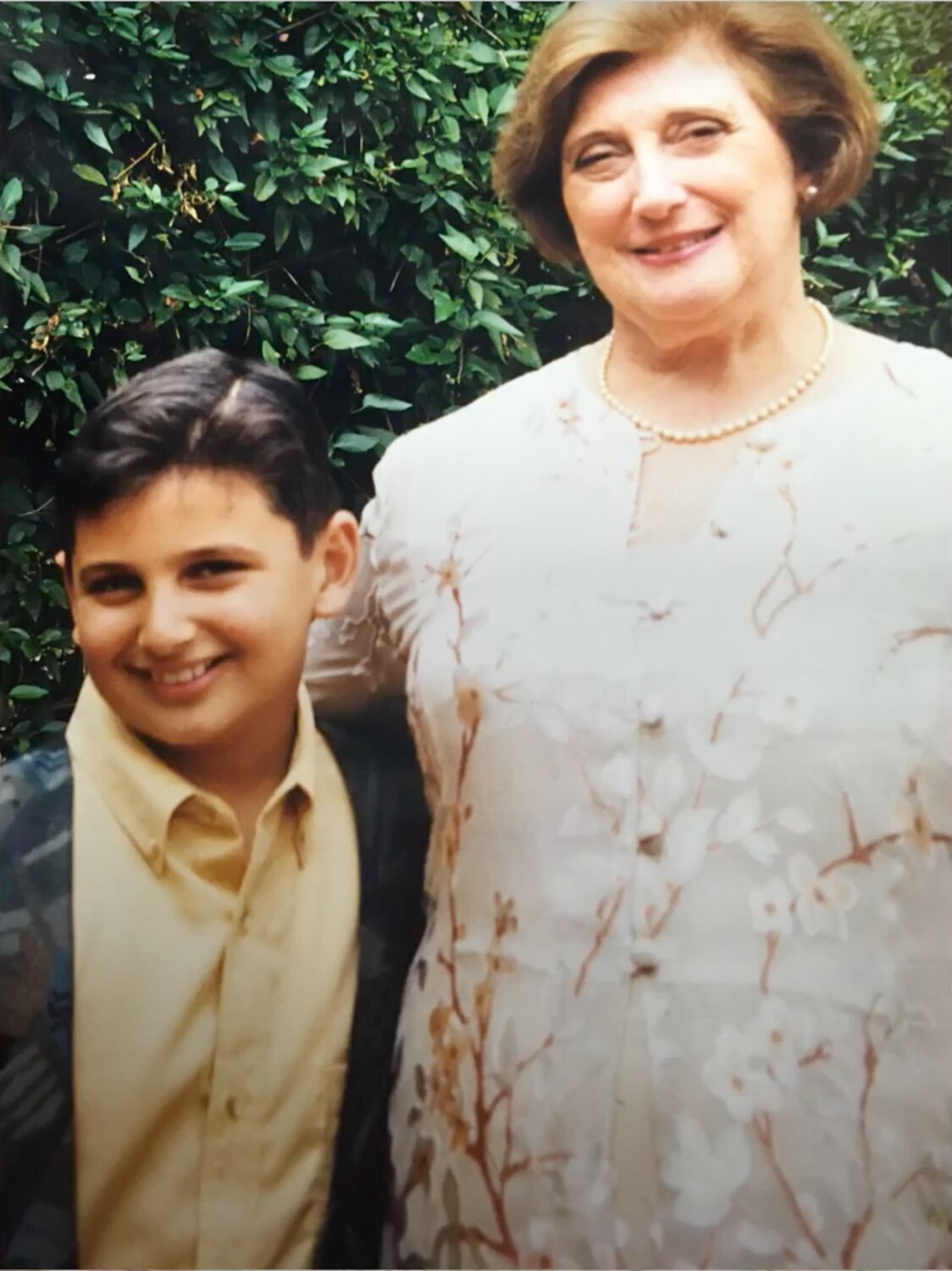 a woman with a cream floral dress and wearing a string of pearls has her arm around a young boy wearing a yellow shirt.