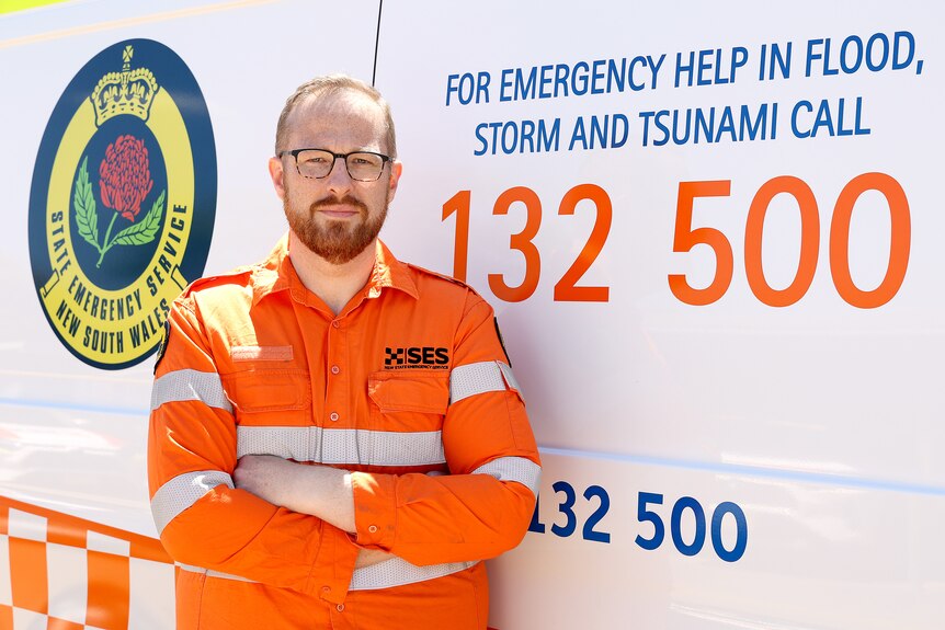 A balding man in SES uniform in front of truck with the SES logo and the emergency help number on it.