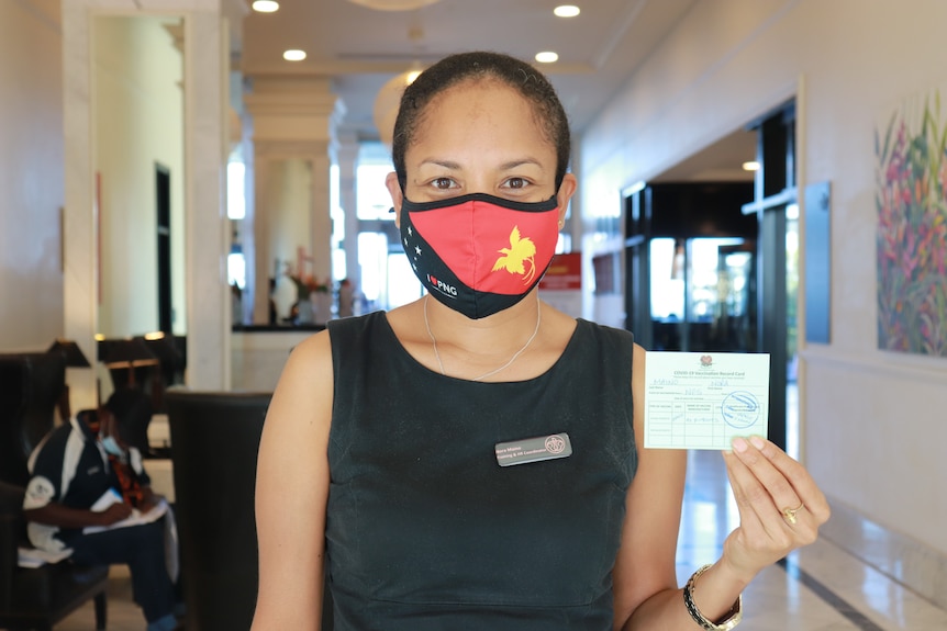 A woman wearing a facemasks with the PNG flag on it shows a card in her left hand and she stands in the lobby of a hotel.