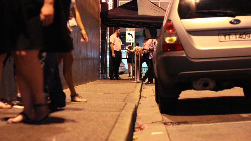 A group of bouncers stand next to a red carpet and rope sectioning off a nightclub in an alleyway.