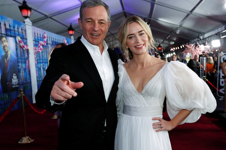 Bob Iger points at the camera wearing a suit while standing next to Emily Blunt in a white puff dress on the red carpet.