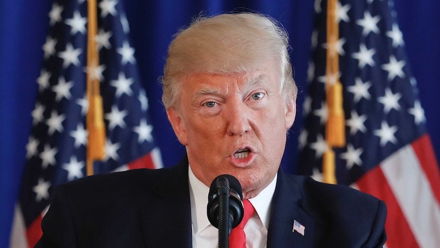 A tight shot of Donald Trump's head and shoulders as he speaks at a lectern, in front of two US flags. His tie is red.