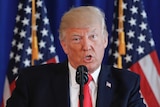 A tight shot of Donald Trump's head and shoulders as he speaks at a lectern, in front of two US flags. His tie is red.