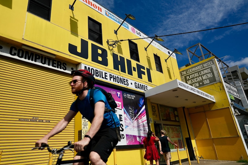A bright yellow JB Hi-Fi store with a cyclist riding past.
