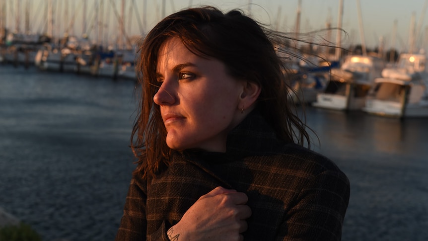 Abbe May stands on the dock of a bay, wearing a jacket, staring into the distance