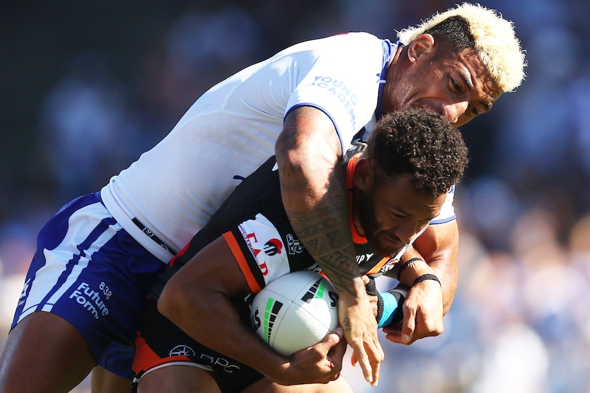 A Wests Tigers NRL player holds the ball while being tackled by a Canterbury opponent.