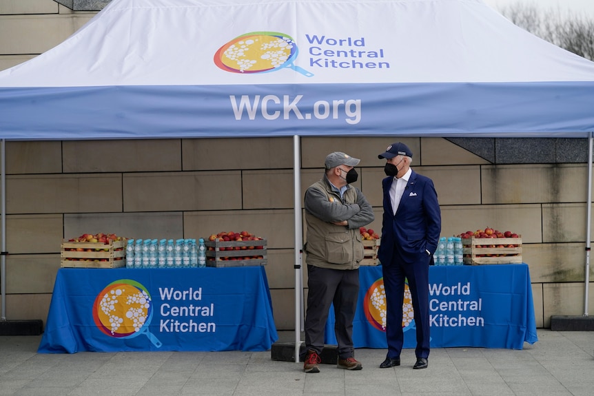 Jose Andres and Joe Biden stand under a tent marked 'World Central Kitchen', in front of benches of food. Both wear masks.