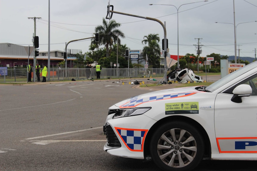 About four police in high-visibility jackets stand around the scene of a car crash, with a police car in the foreground