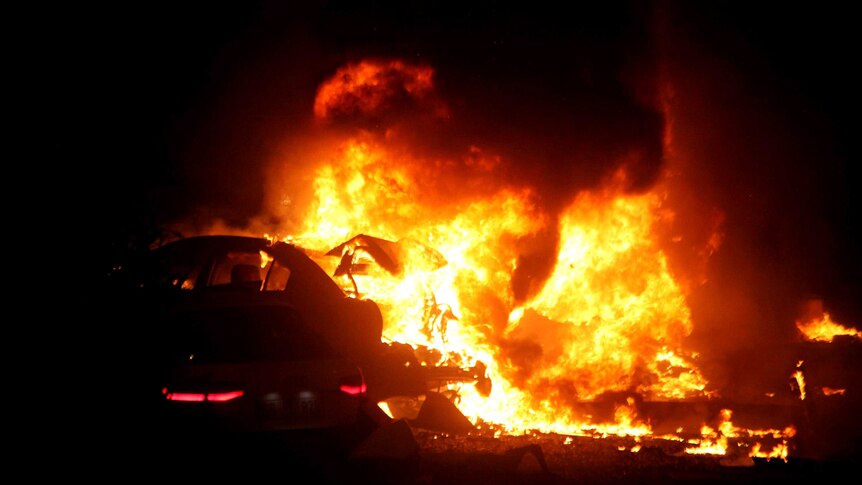 Night photo of bright orange flames pouring from the shell of a car after the explosion.