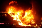 Night photo of bright orange flames pouring from the shell of a car after the explosion.