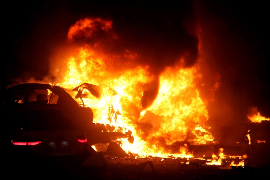 Night photo of bright orange flames pouring from the shell of a car after the explosion.