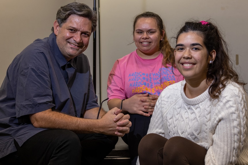 50-year-old indigenous man with salt-and-pepper short hair sits with two young Indigenous women, all smiling.