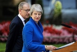 Theresa May looks past the camera in an electric blue blazer in a wooden lectern with her partner Philip beside her.
