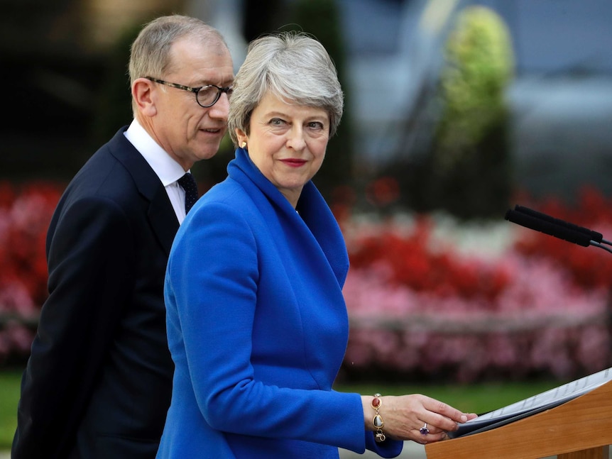 Theresa May looks past the camera in an electric blue blazer in a wooden lectern with her partner Philip beside her.