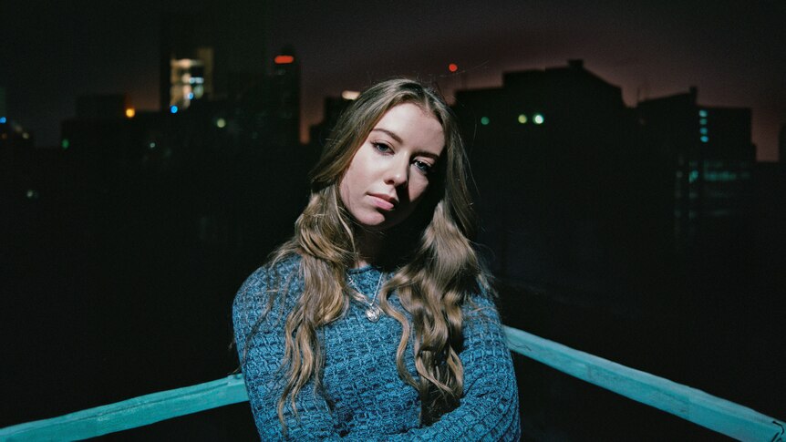 Darcie Haven stands against a blue railing in the dark wearing a blue jumper and her brown hair out.