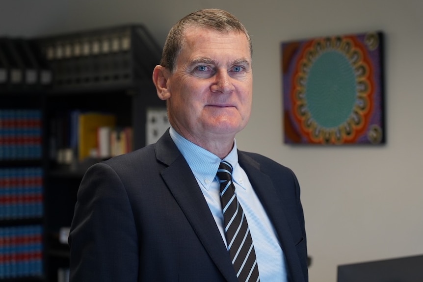 A slightly smiling middle-aged man with short dark blond hair, wearing a suit and tie, stood in front of a bookshelf, drawing pictures.