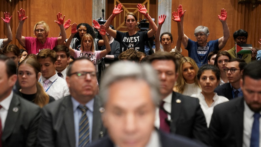 Antony Blinken is seen out of focus as protesters behind him lift their hands with red-painted palms