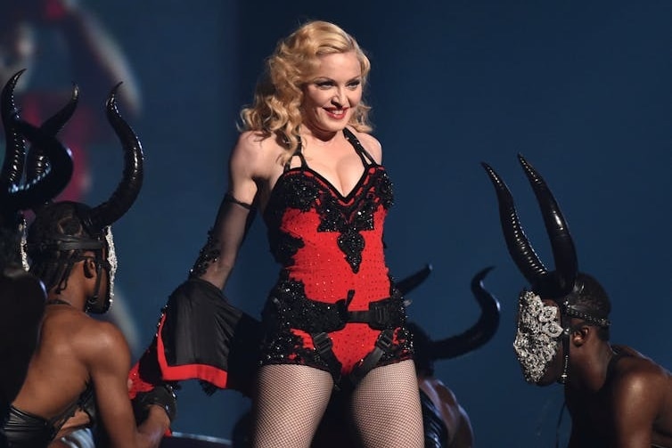 Madonna performs on stage in a red corset, with horned dancers around her