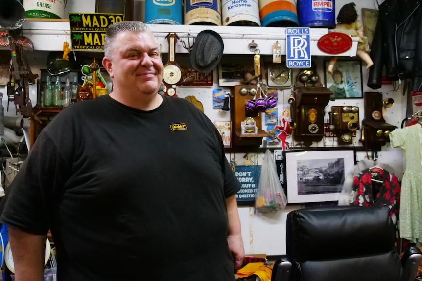 A man in a black shirt stands in a quaint shop crammed with oddities.