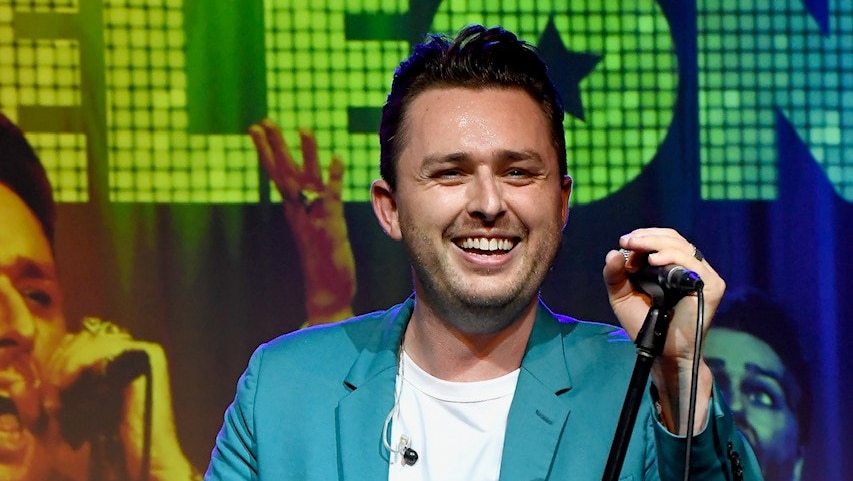 A man in a light blue suit stands on a stage in front of coloured lights, holding a microphone and smiling.