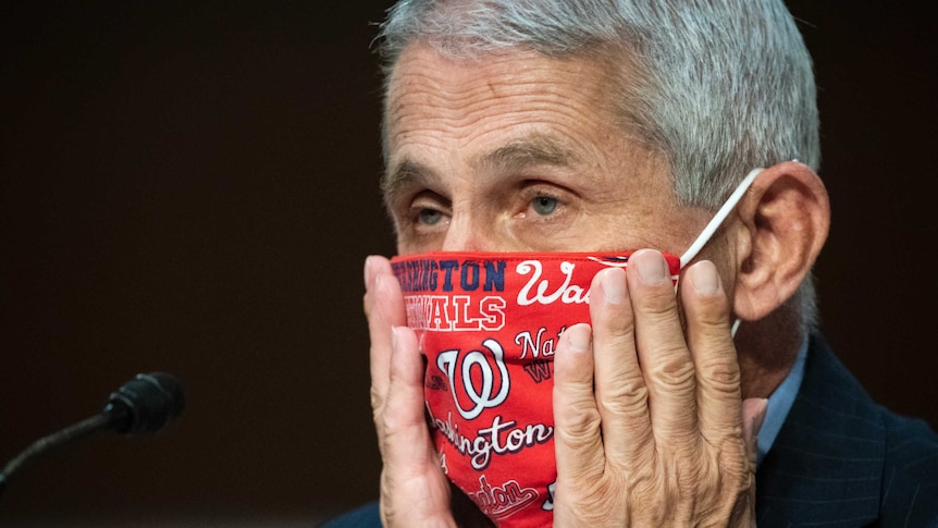 An elderly man with short grey hair places his palms on his hands on his cheeks and adjusts a red face mask.