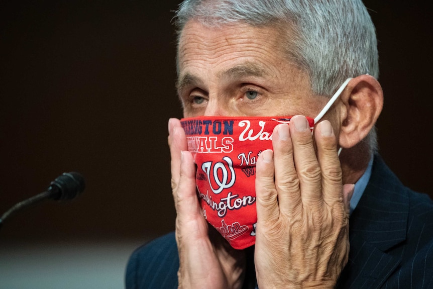 An elderly man with short grey hair places his palms on his hands on his cheeks and adjusts a red face mask.