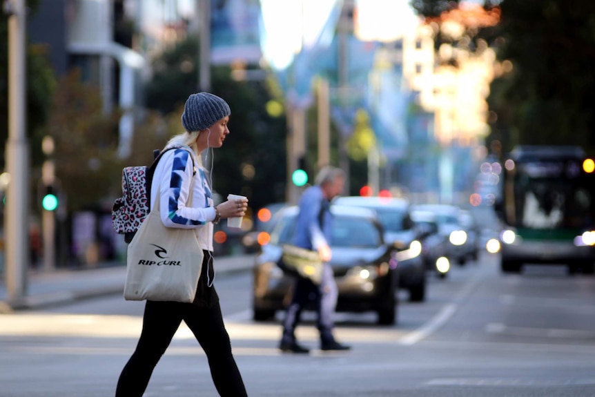 A woman crosses the road carrying a cup of coffee and a bag on St Georges Tce in Perth.