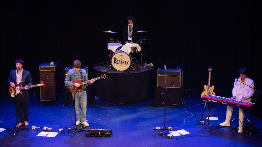 Four men in a band on stage playing tribute to The Beatles.