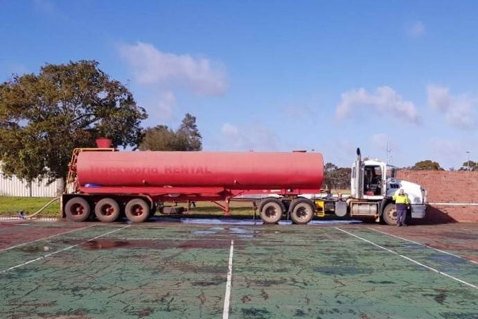 A red tanker on a water haulage truck parked on an old tennis court with a hose extending towards the camera.