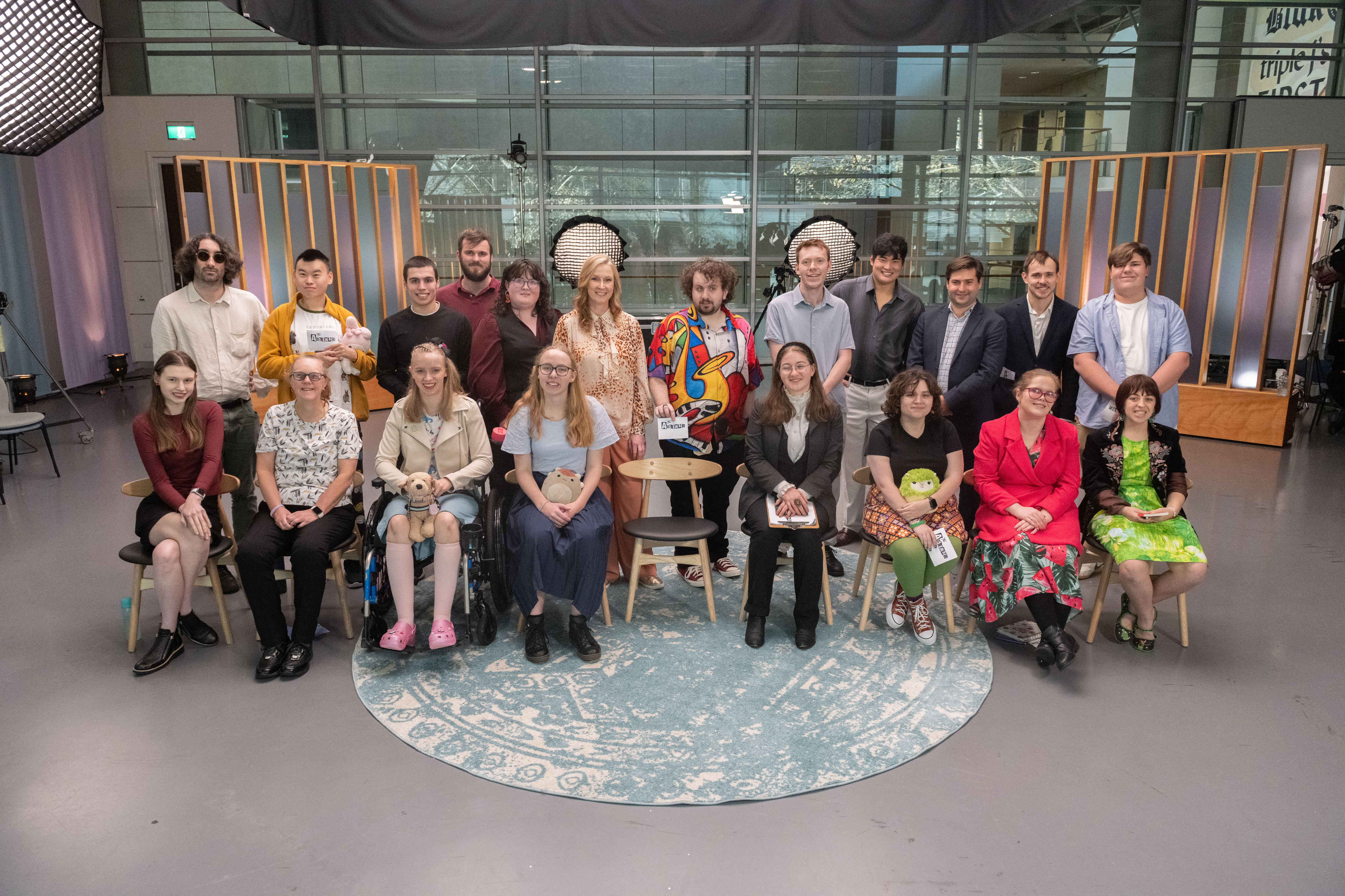 Um grupo de 20 estudantes sorridentes está em um estúdio com a apresentadora Leigh Sales no centro. 
