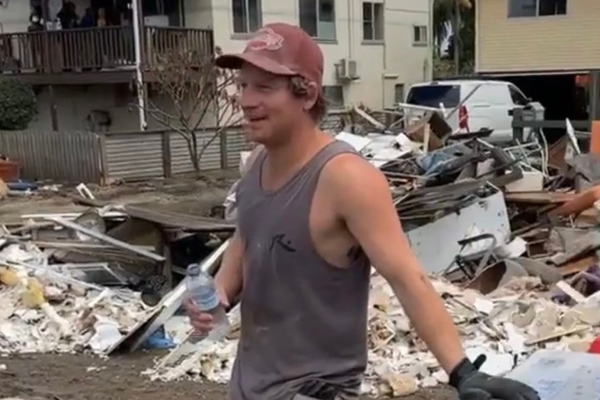 man with cap in singlet leans on trailer with water bottle in hand in front of debris 