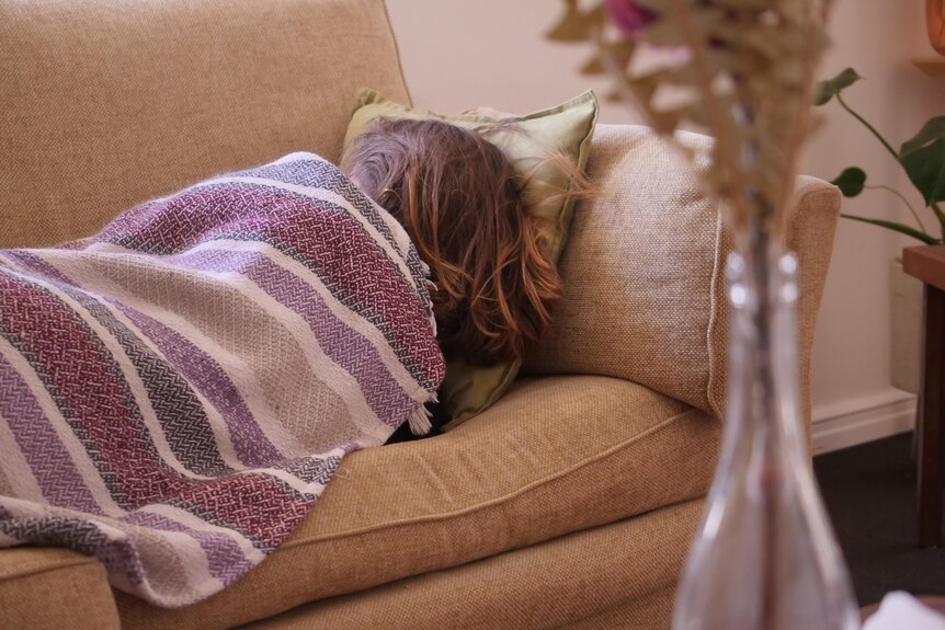 A young girl is curled on a couch under a blanket, facing away from the camera