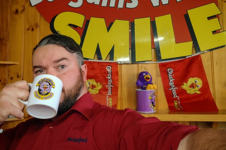 A man in a red shirt that says Chickenfeed is drinking from a Chickenfeed branded mug with Chickenfeed flags in background