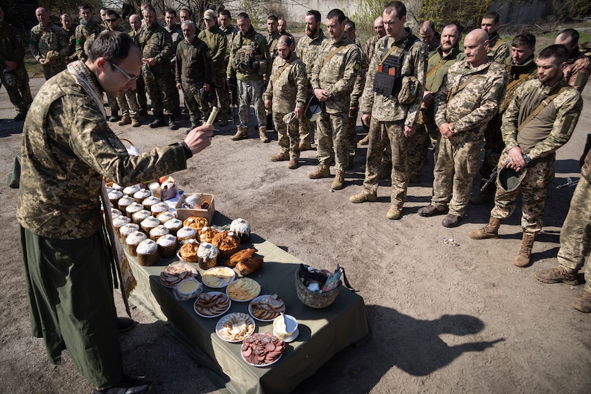 A military Orthodox priest blesses traditional food while a row of soldiers look on.