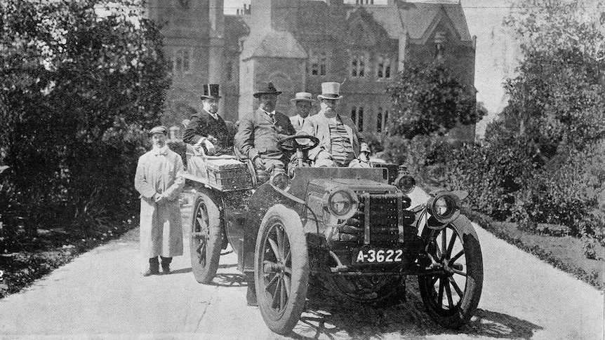 old black and white photo of five men in an old motor car, wearing top hats, in front of an old sandstone building