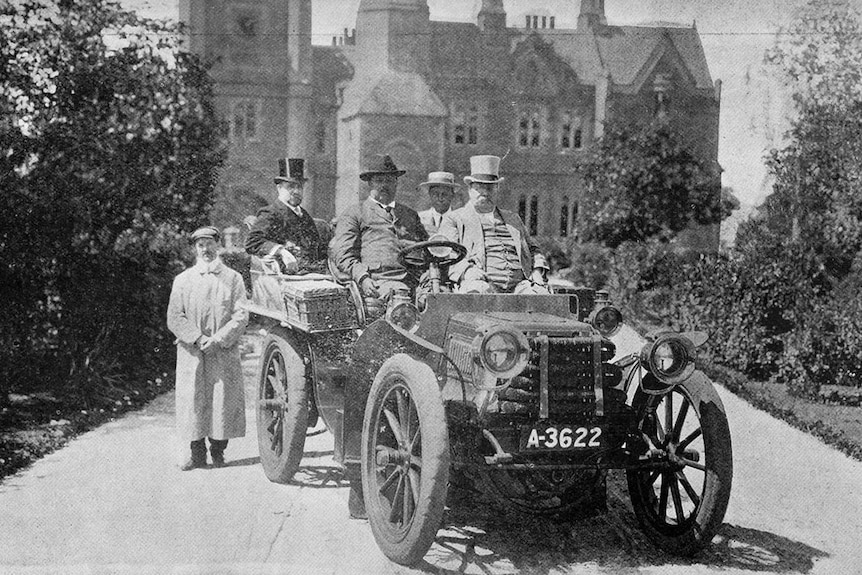old black and white photo of five men in an old motor car, wearing top hats, in front of an old sandstone building