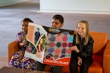 Three children hold a big book with Aboriginal design on its cover