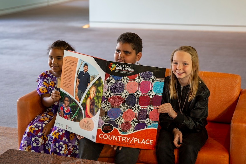 Three children hold a big book with Aboriginal design on its cover