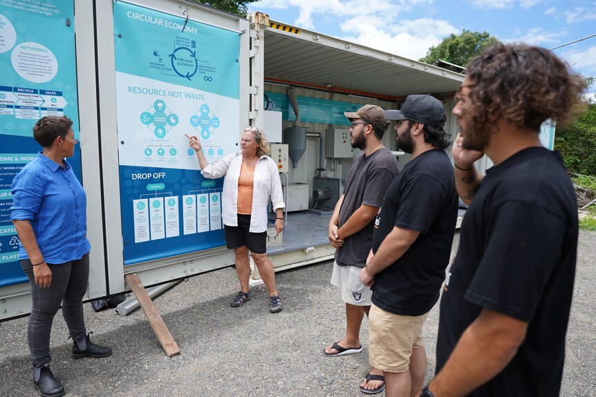 Woman pointing at a poster about recycling. 3 men and a woman dressed casually looking on and listening. 