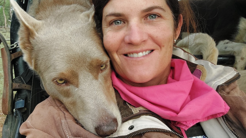 a woman in a pink scarf smiles, her dog rests his head on her right shoulder
