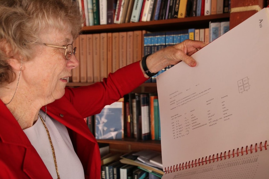 Cheryl Praeger stands in front of a bookcase holding a mathematics book.