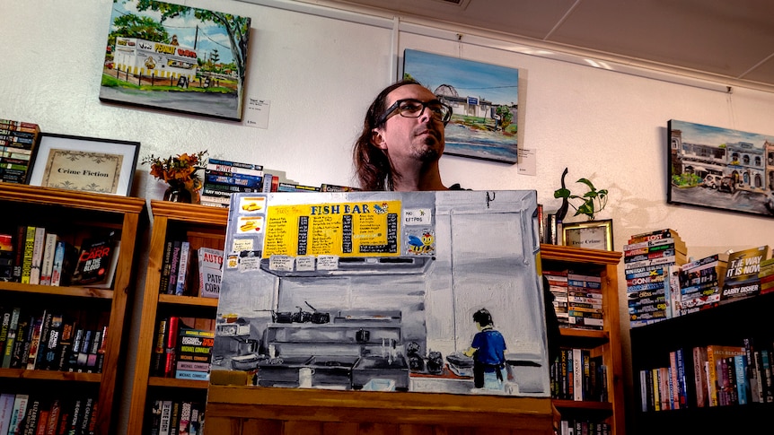 A man holds a painting of a fish and chip shop worker in a book shop setting.